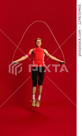 Fit young man in red training top jumping with skipping rope mid-air, showing focus and agility in active motion against red background. Fit young man in red training top jumping with skipping rope mid-air, showing focus and agility in active motion against red background. 124650468