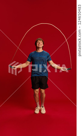 Dynamic photo of young man doing high-speed jump rope workout against red background, expressing joy and movement. 124650483
