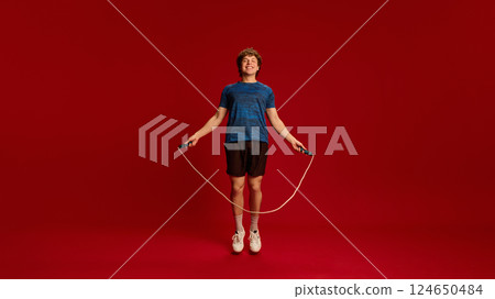 Energetic man mid-air skipping rope, smiling during dynamic cardio session against red studio background. Energetic man mid-air skipping rope, smiling during dynamic cardio session against red studio background. 124650484