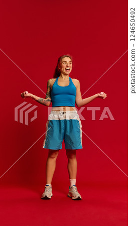 Full body photo of slim young woman dressed blue uniform with joyful expression, doing biceps curls in motion against red studio background. Full body photo of slim young woman dressed blue uniform with joyful expression, doing biceps curls in motion against red studio background. 124650492