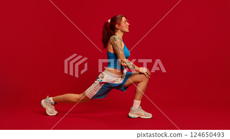 Side view, energetic photo of focused on training young woman dressed blue uniform, doing alternating forward lunge against red background. Side view, energetic photo of focused on training young woman dressed blue uniform, doing alternating forward lunge against red background. 124650493