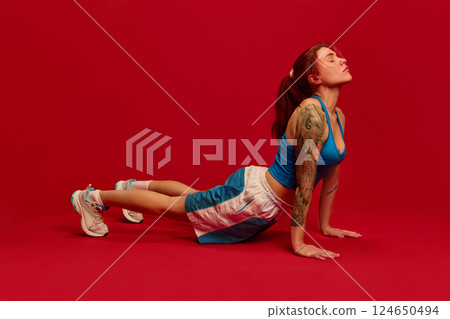 Full length photo of young woman dressed blue sports attire, stretching back, standing in yoga pose Upward-Facing Dog against red background. Full length photo of young woman dressed blue sports attire, stretching back, standing in yoga pose Upward-Facing Dog against red background. 124650494