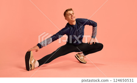 Sporty man smiling while stretching legs sideways, reaching for foot on pink background, wearing striped sleeve sportswear. 124650531