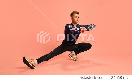 Flexible athlete performing side lunge with arms crossed, dressed in sporty outfit and sneakers, demonstrating dynamic stretch against pink background. 124650532