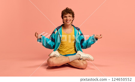 Smiling man in vintage sportswear sitting cross-legged, practicing meditation with relaxed posture against pink background. 124650552
