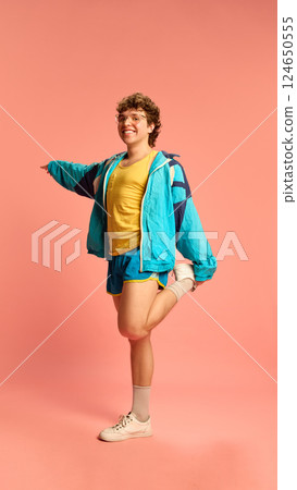Cheerful man in retro sportswear stretching quadriceps, balancing on one leg with hand out against soft pink background. 124650555