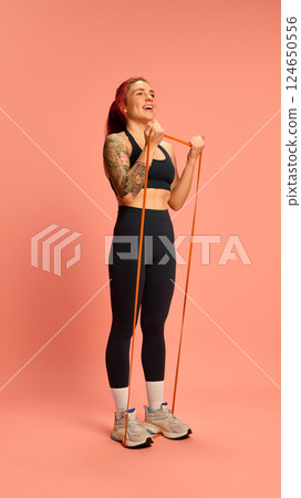 Energetic woman in black workout set standing on resistance band, curling arms with effort and joy against soft pink background. 124650556