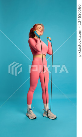 Young woman in pink activewear exercising with resistance band, doing bicep curls, showing muscle engagement and positive attitude. Young woman in pink activewear exercising with resistance band, doing bicep curls, showing muscle engagement and positive attitude. 124650560