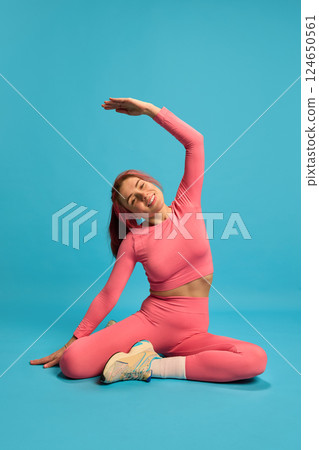 Young sporty woman stretching sideways on floor in pink activewear, smiling and showing flexibility against blue studio background. Young sporty woman stretching sideways on floor in pink activewear, smiling and showing flexibility against blue studio background. 124650561