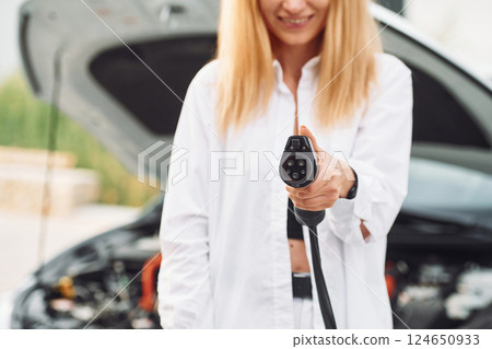 Holding charger. Young woman in white clothes is with her electric car at daytime 124650933