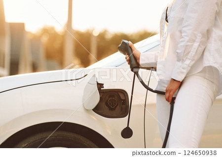 Close up view. Holding charger. Young woman in white clothes is with her electric car at daytime 124650938