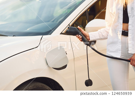 Close up view. Holding charger. Young woman in white clothes is with her electric car at daytime Close up view. Holding charger. Young woman in white clothes is with her electric car at daytime 124650939