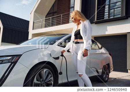 Charging process. Young woman in white clothes is with her electric car at daytime Charging process. Young woman in white clothes is with her electric car at daytime 124650940