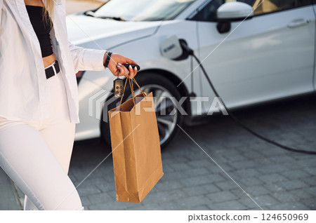 With sopping bag. Young woman in white clothes is with her electric car at daytime With sopping bag. Young woman in white clothes is with her electric car at daytime 124650969