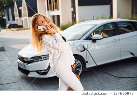 Holding phone and talking by it. Young woman in white clothes is with her electric car at daytime Holding phone and talking by it. Young woman in white clothes is with her electric car at daytime 124650972