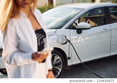 Young woman in white clothes is with her electric car at daytime Young woman in white clothes is with her electric car at daytime 124650975