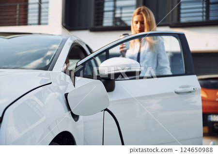 Opening the door. Young woman in white clothes is with her electric car at daytime 124650988