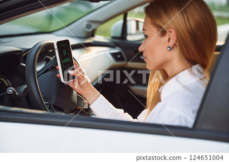 Sitting inside with smartphone. Young woman in white clothes is with her electric car at daytime 124651004