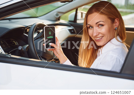 Sitting inside with smartphone. Young woman in white clothes is with her electric car at daytime 124651007