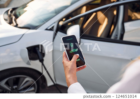 Smartphone monitoring of charging. Young woman in white clothes is with her electric car at daytime 124651014