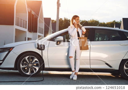 Holding shopping bag. Young woman in white clothes is with her electric car at daytime 124651018