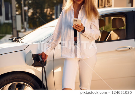 Illuminated by sunlight. Young woman in white clothes is with her electric car at daytime Illuminated by sunlight. Young woman in white clothes is with her electric car at daytime 124651020