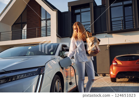 Holding shopping bag. Young woman in white clothes is with her electric car at daytime Holding shopping bag. Young woman in white clothes is with her electric car at daytime 124651023