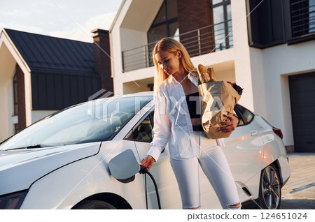 Holding shopping bag. Young woman in white clothes is with her electric car at daytime 124651024