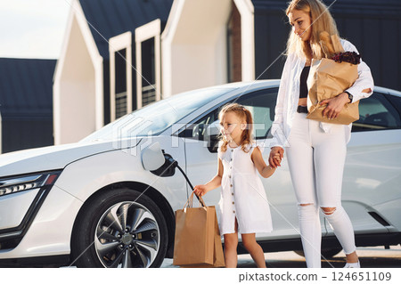 With shopping bags. Young woman with her little daughter is with their electric car outdoors With shopping bags. Young woman with her little daughter is with their electric car outdoors 124651109