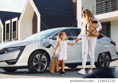 With shopping bags. Young woman with her little daughter is with their electric car outdoors With shopping bags. Young woman with her little daughter is with their electric car outdoors 124651110