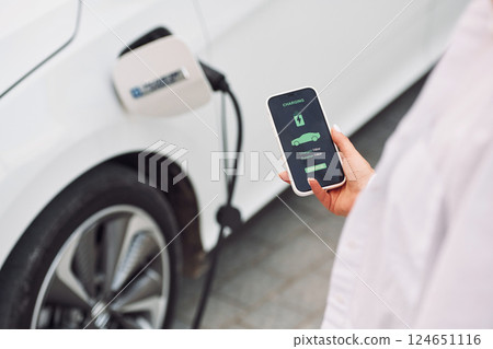Close up view. Woman charging her electric car and monitoring process on smartphone Close up view. Woman charging her electric car and monitoring process on smartphone 124651116