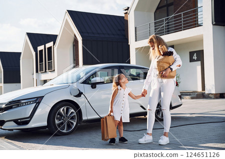 Standing together. Young woman with her little daughter is with their electric car outdoors 124651126