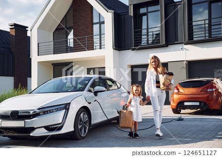 Holding bags while vehicle is on a charge. Young woman with her little daughter is with their electric car outdoors 124651127