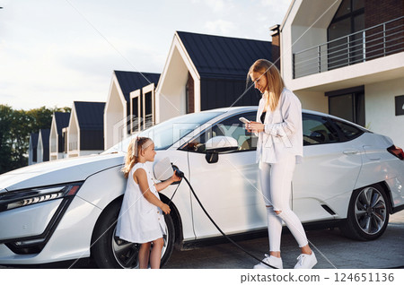 Charging the vehicle together. Young woman with her little daughter is with their electric car outdoors Charging the vehicle together. Young woman with her little daughter is with their electric car outdoors 124651136