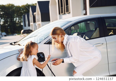 Charging the vehicle together. Young woman with her little daughter is with their electric car outdoors Charging the vehicle together. Young woman with her little daughter is with their electric car outdoors 124651137