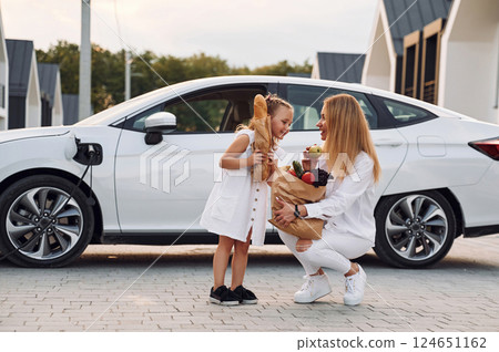 Holding bag with products. Young woman with her little daughter is with their electric car outdoors 124651162