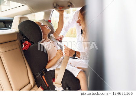 Girl is in a special baby seat. Young woman with her little daughter is with their electric car outdoors Girl is in a special baby seat. Young woman with her little daughter is with their electric car outdoors 124651166
