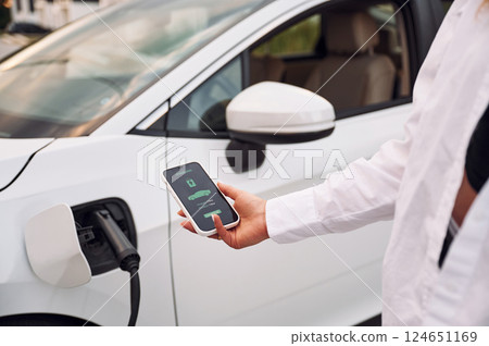 Close up view. Woman charging her electric car and monitoring process on smartphone Close up view. Woman charging her electric car and monitoring process on smartphone 124651169