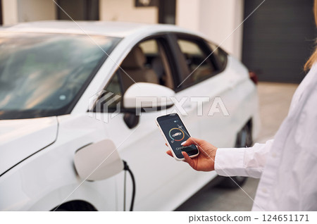 Near garage. Close up view. Woman charging her electric car and monitoring process on smartphone Near garage. Close up view. Woman charging her electric car and monitoring process on smartphone 124651171