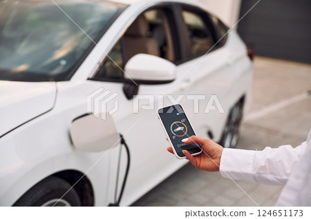 Near garage. Close up view. Woman charging her electric car and monitoring process on smartphone Near garage. Close up view. Woman charging her electric car and monitoring process on smartphone 124651173