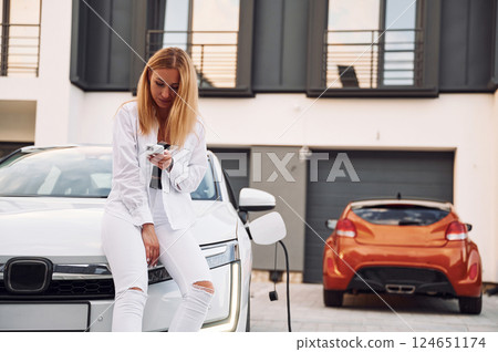 Near the home. Young woman in white clothes is with her electric car at daytime Near the home. Young woman in white clothes is with her electric car at daytime 124651174