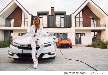 Near the home. Young woman in white clothes is with her electric car at daytime 124651175