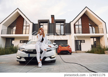 Near the home. Young woman in white clothes is with her electric car at daytime Near the home. Young woman in white clothes is with her electric car at daytime 124651176