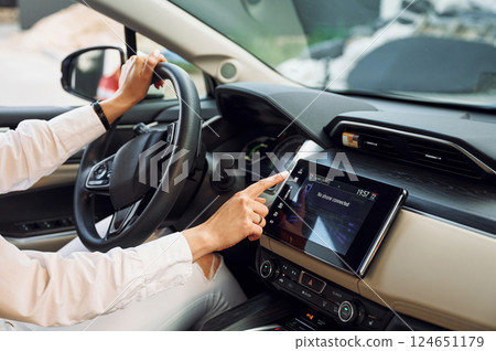 Navigating the vehicle's interface. Young woman in white clothes is with her electric car at daytime Navigating the vehicle's interface. Young woman in white clothes is with her electric car at daytime 124651179