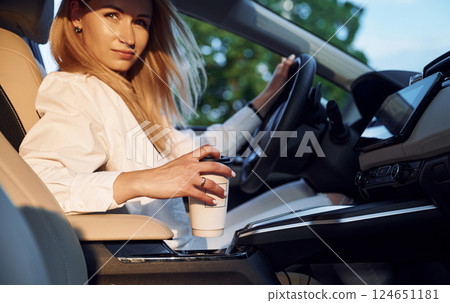 With cup of drink. Young woman in white clothes is with her electric car at daytime With cup of drink. Young woman in white clothes is with her electric car at daytime 124651181