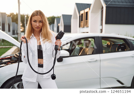Standing and holding charging cable. Young woman in white clothes is with her electric car at daytime Standing and holding charging cable. Young woman in white clothes is with her electric car at daytime 124651194
