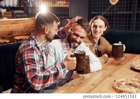 Smiling, knocking mugs, pizza on the table. Three friends are in the bar drinking beer and having a good time Smiling, knocking mugs, pizza on the table. Three friends are in the bar drinking beer and having a good time 124651581