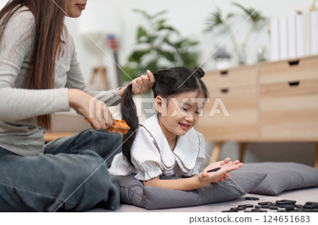 Care and Connection. A mother lovingly brushes her daughter's hair while she enjoys playing with dominoes. 124651683