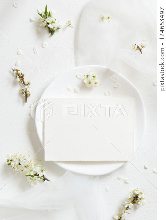 Blank card on plate near tulle and wild plums white flowers top view, romantic wedding mockup 124653497