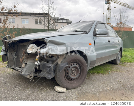 A silver car with severe front-end damage is parked near a construction site, showing a crushed fender, missing bumper 124655078